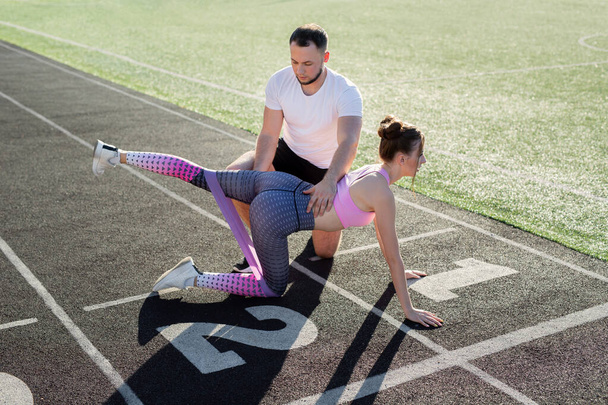 Coach plays sports with a young girl at the stadium. Leg exercise with elastic band. - 写真・画像