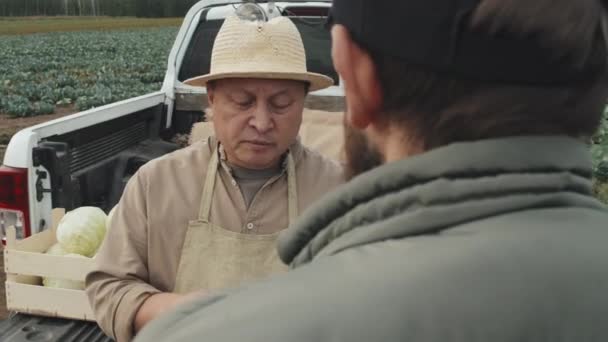 Πλάνα πάνω από τον ώμο ενός ώριμου Ασιάτη αγρότη που δίνει οδηγίες στον νεαρό άνδρα για την παράδοση λαχανικών σε πελάτες - Πλάνα, βίντεο