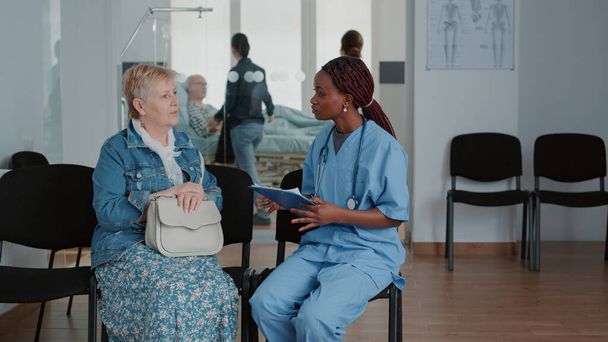 Primer plano de la enfermera afroamericana hablando con el paciente mayor en la sala de espera - Foto, Imagen
