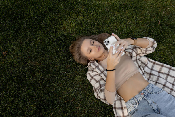 Genç kadın yeşil çimlere uzanıyor ve telefona bakarken gülümsüyor. - Fotoğraf, Görsel