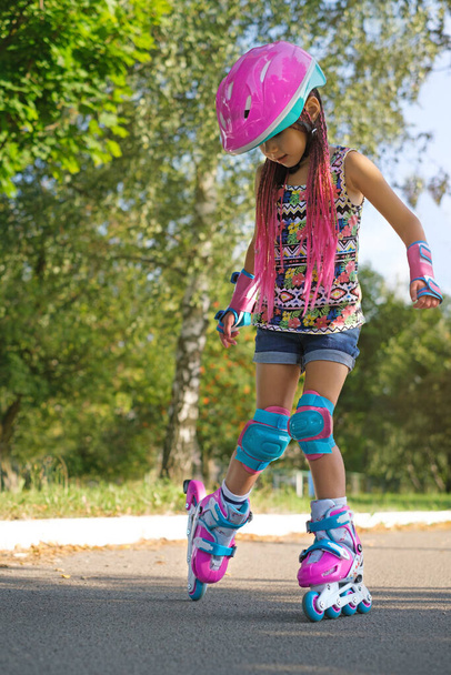 Menina com rosa afro tranças tranças dreadlocks em um capacete, cotovelos protetores e joelheiras passeios canyons rolo na rua na cidade em caminhos asfaltados. Kit de proteção para patinadores infantis - Foto, Imagem