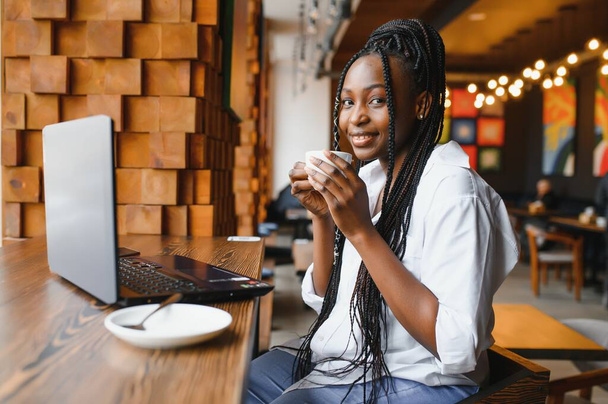 Een jong Afro-Amerikaans meisje zit in een restaurant en typt op haar laptop. mooi meisje werken op computer in cafe. - Foto, afbeelding