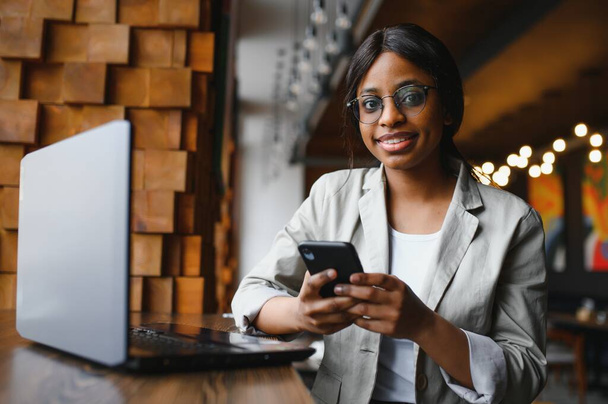 Head shot portrait of happy smiling African American woman sitting at table in cafe, looking at camera, excited female posing, working at computer, doing homework, preparing report in coffee house - 写真・画像