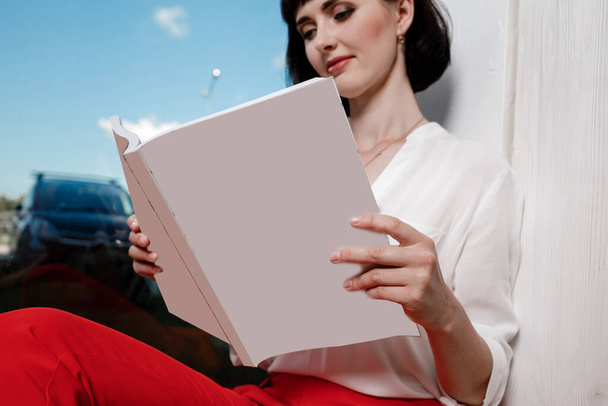 Piękna młoda kobieta trzymająca magazyn z makietą. dziewczyna w białej koszuli i czerwonych spodniach siedzi w pobliżu okna. - Zdjęcie, obraz