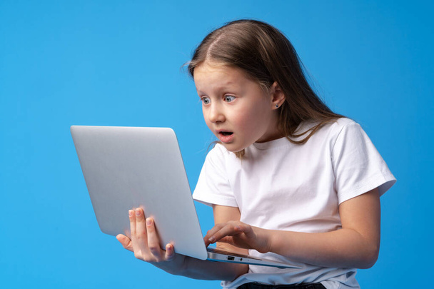 Portrait of a little girl holding laptop computer while standing against blue background - Foto, Imagem