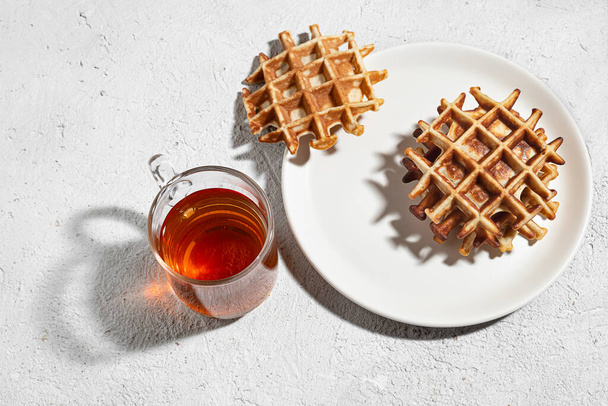 Gofres belgas en un plato blanco y una taza de té en una mesa ligera, sabroso concepto de desayuno, vista superior - Foto, imagen