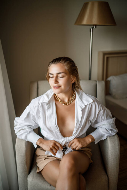 Hermosa mujer atractiva con el pelo rubio se sienta en una habitación en una silla con una camisa blanca y posando maravillosamente - Foto, Imagen
