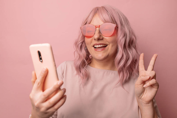 Attractive young woman wears pink sunglasses makes v sign shows peace gesture has glad expression uses smartphone isolated over pink wall background - Photo, Image