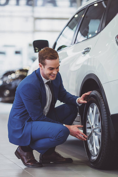 Przystojny młody biznesmen w klasycznym niebieskim garniturze uśmiecha się podczas badania samochodu na pokazie motoryzacyjnym - Zdjęcie, obraz