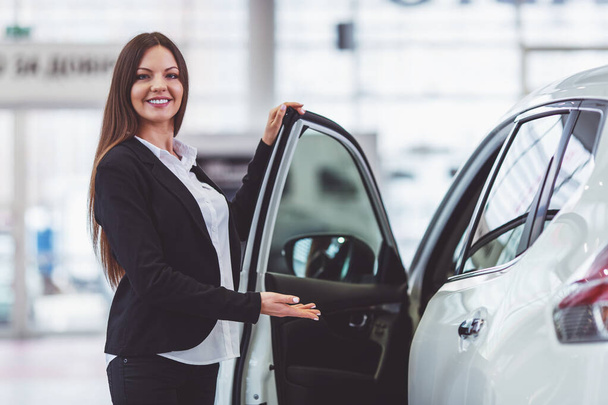 schöne junge Frau im klassischen Anzug lächelt und blickt in die Kamera, während sie ein Auto auf einer Automesse präsentiert - Foto, Bild