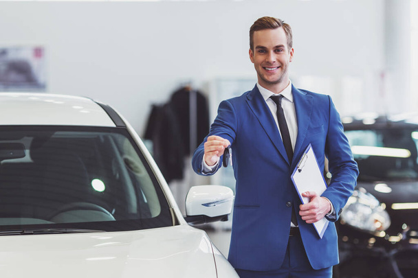 Knappe jonge man in klassieke blauwe pak is glimlachend, camera kijken en het aanbieden van autosleutels tijdens de presentatie van de auto in een motor show - Foto, afbeelding