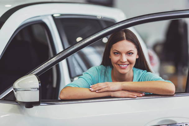 Belle jeune femme en vêtements décontractés sourit et regarde la caméra tout en s'appuyant sur une nouvelle voiture dans un salon de l'automobile
 - Photo, image