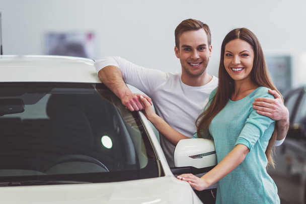 Beau jeune couple en vêtements décontractés sourit et regarde la caméra tout en s'appuyant sur leur nouvelle voiture dans un salon de l'automobile
 - Photo, image