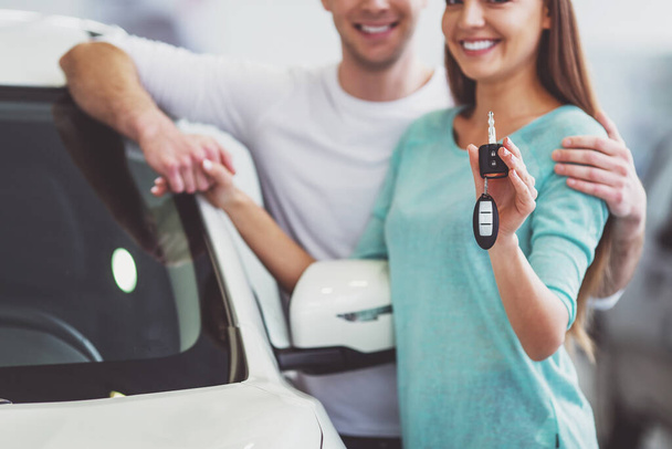 Bela jovem casal está sorrindo e olhando para a câmera enquanto se inclina em seu novo carro em um show de motor. Mulher está segurando chaves do carro, close-up
 - Foto, Imagem