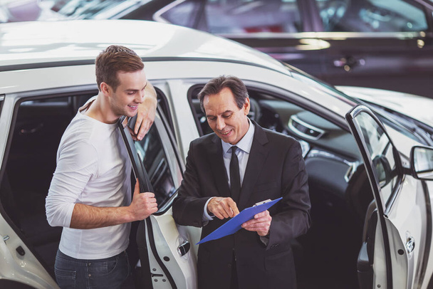 Bello venditore di mezza età sta presentando una macchina al bel giovane cliente in un salone automobilistico
 - Foto, immagini