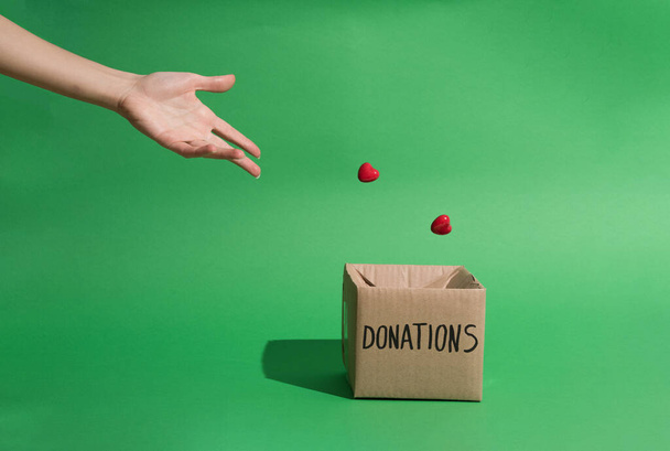 Schwebende winzige Herzen, die von Hand geworfen werden, fallen in Spendenbox für wohltätige Zwecke vor grünem Hintergrund - Foto, Bild