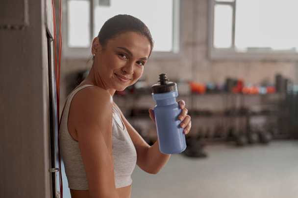 Kobieta w sportowym stroju w klubie sportowym, uśmiechnięta i trzymająca butelkę wody - Zdjęcie, obraz