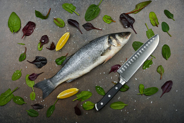 Close up one fresh raw European sea bass fish on table, with kitchen knife, lemon wedges, and salad leaves, elevated top view, directly above - 写真・画像