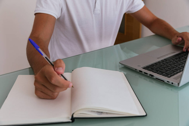 Oldalkép egy felismerhetetlen fiatalemberről fehér pólóban, aki otthon dolgozik a számítógépénél, és jegyzetel egy jegyzetfüzetben. - Fotó, kép