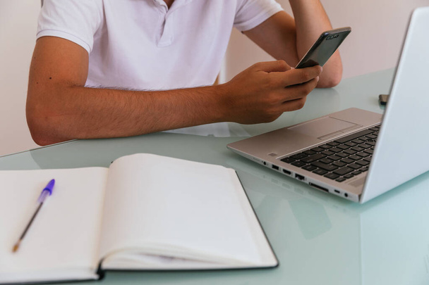 Бічна фотографія невпізнаваного молодого чоловіка в білій сорочці поло, що працює вдома зі смартфоном у руці, ноутбуком та блокнотом
 - Фото, зображення