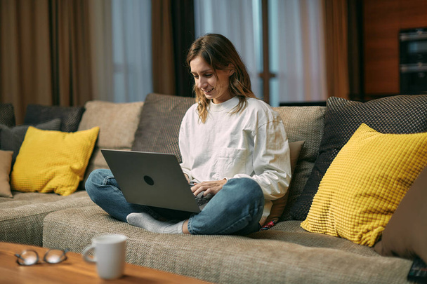 Happy young creative girl freelancer working on laptop sitting on sofa in living room at home. Remote distant online job - Фото, зображення