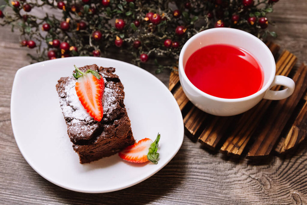 Une portion de brownie au chocolat et une tasse de thé aux fruits pour prendre le temps de faire une pause. Sur un plateau en bois, une tasse de thé chaud. Baies artificielles sauvages sur un fond. - Photo, image