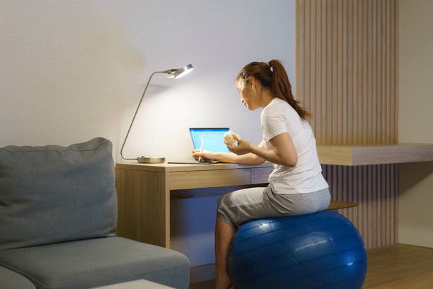 Asian woman relaxing on yoga ball and eating sandwich and chatting online with team of colleagues on laptop while work from home at home at night. - Fotografie, Obrázek