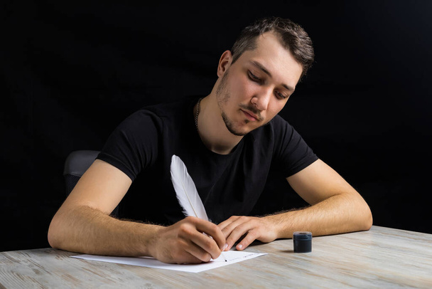 Młody przystojny i poważny mężczyzna ubrany w pióro czarny pisze na białym arkuszu papieru na czarnym tle w niskim kluczu. - Zdjęcie, obraz