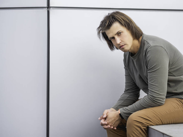 Millennial man with long hair and tense look sits near grey wall. Serious man in earth toned casual clothes is staring in camera. Copy space. - Photo, Image
