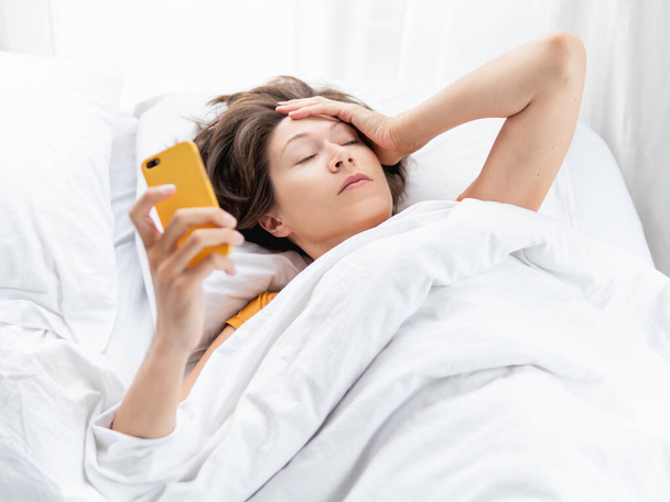 Sleepy woman looks on smartphone screen. Checking e-mail box right after waking up. Using wireless technology in bed. - Fotó, kép