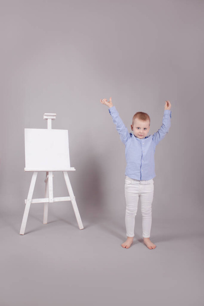 μικρό αγόρι στέκεται κοντά στον πίνακα παρουσίασης καβαλέτο με λευκό καμβά σετ. Απομονωμένο σε γκρι φόντο. Κενό μακιγιάρισμα - Φωτογραφία, εικόνα