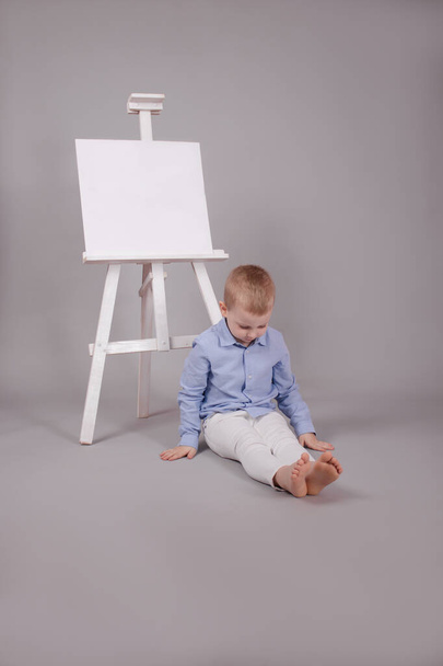 Menino de pé perto do cavalete da mesa de apresentação com conjunto de lona em branco. Isolado em fundo cinza. Mochila em branco - Foto, Imagem