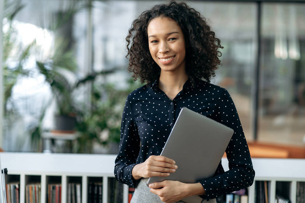Retrato de bonita, bem sucedida afro-americana jovem empresária, chefe de departamento, gerente de topo, com um laptop, de pé em um escritório moderno, olhando para a câmera, sorrindo amigável - Foto, Imagem