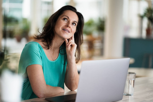 thoughtful brooding remote working dark haired woman sitting infront of a laptop or notebook in casual outfit on her work desk in her modern airy bright living room home office with many windows - Foto, Bild