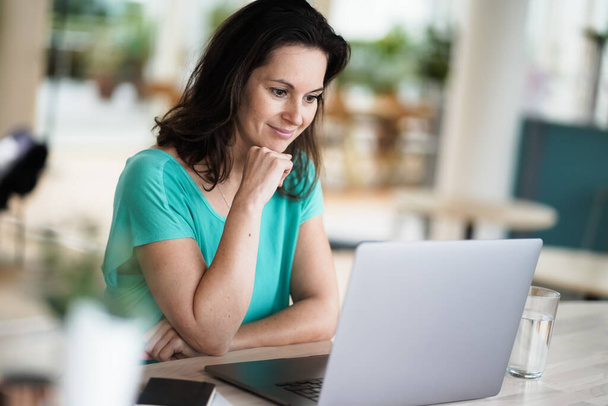 thoughtful brooding remote working dark haired woman sitting infront of a laptop or notebook in casual outfit on her work desk in her modern airy bright living room home office with many windows - Foto, Imagen