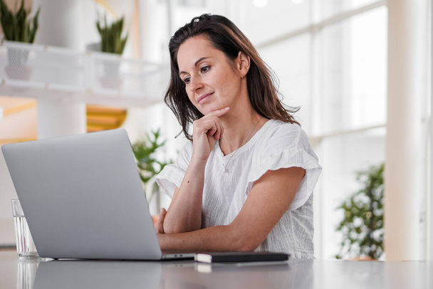 thoughtful brooding remote working dark haired woman sitting infront of a laptop or notebook in casual outfit on her work desk in her modern airy bright living room home office with many windows - Fotografie, Obrázek