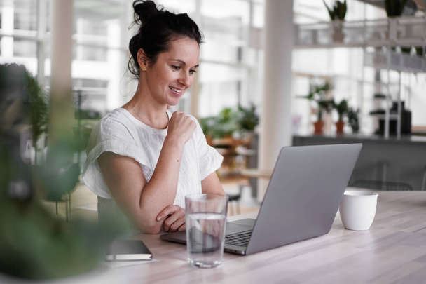 happy smiling  successful remote working dark haired woman cheerful infront of laptop or notebook in casual outfit sitting on work desk in her modern loft living room home office having a video chat - Foto, Bild