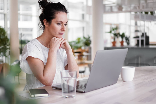 thoughtful brooding remote working dark haired woman sitting infront of a laptop or notebook in casual outfit on her work desk in her modern airy bright living room home office with many windows - Fotó, kép