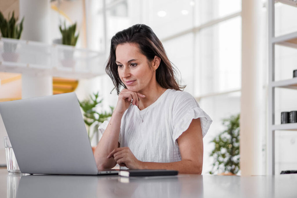 thoughtful brooding remote working dark haired woman sitting infront of a laptop or notebook in casual outfit on her work desk in her modern airy bright living room home office with many windows - Fotoğraf, Görsel