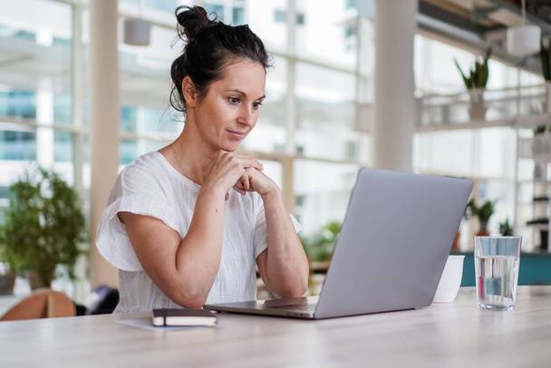 átgondolt tűnődő távoli dolgozó sötét hajú nő ül infravörös egy laptop vagy notebook alkalmi ruhát a munkaasztalán az ő modern levegős világos nappali otthoni iroda sok ablakot - Fotó, kép