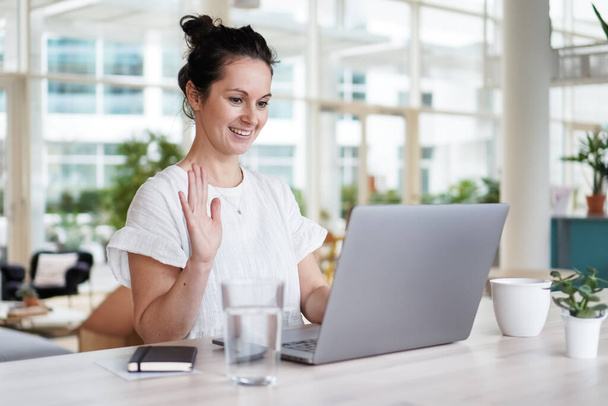 happy smiling  successful remote working dark haired woman cheerful infront of laptop or notebook in casual outfit sitting on work desk in her modern loft living room home office having a video chat - Foto, imagen