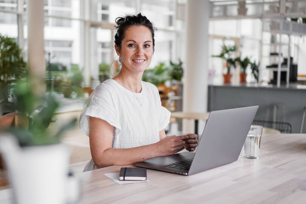 szczęśliwy uśmiechnięty sukces zdalnej pracy ciemnowłosy kobieta wesoły naprzeciwko laptopa lub notebooka w casual strój siedzi na biurku w nowoczesnym poddaszu salon biuro o wideo czat - Zdjęcie, obraz
