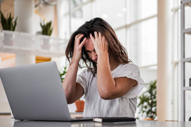 sad depressed remote working woman sitting disappointed with headache infront of a laptop or notebook on her work desk in modern airy bright living room home office with many windows - Photo, Image