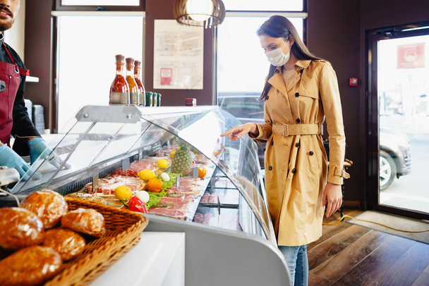 Happy young woman in face mask choosing meat from glass cabinet in grocery store - Photo, image