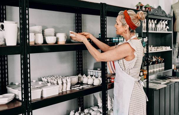 Another addition to the collection. Cropped shot of an attractive mature woman standing and organising her pottery on a shelf in her workshop. - Foto, afbeelding