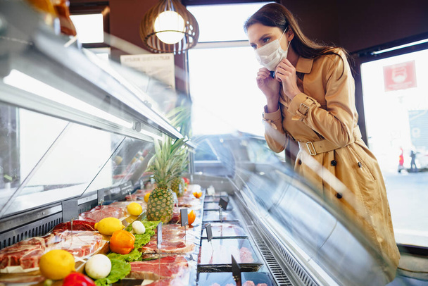 Happy young woman in face mask choosing meat from glass cabinet in grocery store - Foto, afbeelding