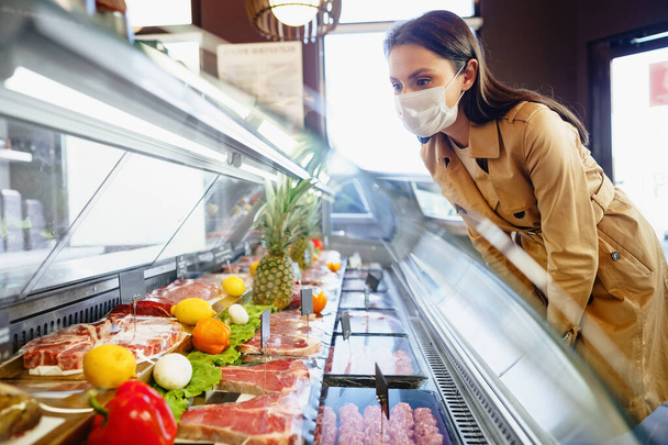 Happy young woman in face mask choosing meat from glass cabinet in grocery store - Fotografie, Obrázek