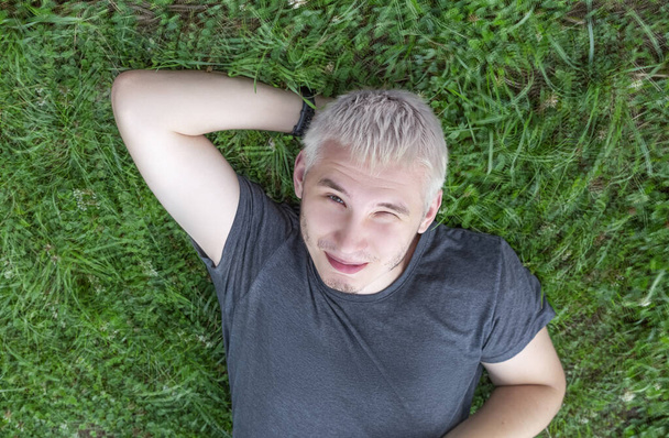 A cheerful guy in a gray T-shirt is lying on a green lawn with one eye squinted - Photo, Image