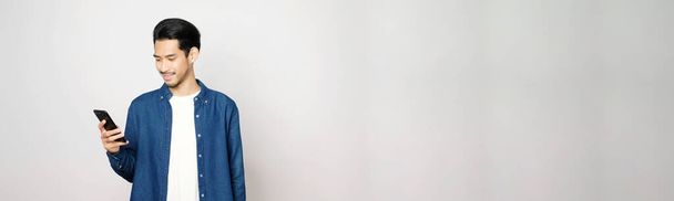 asiatico uomo tenendo cellulare sorridente mentre in piedi isolato su sfondo grigio con copia spazio per il testo, persone su smart phone, tecnologia stile di vita - Foto, immagini
