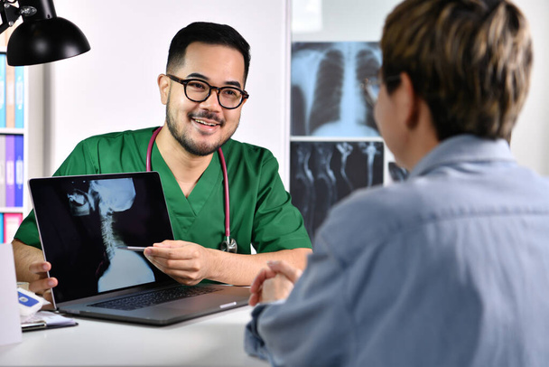 Vriendelijk geruststellend en attent. glimlach jong aziatisch man arts in chirurgische uniform consulting en uitleggen vrouwelijke patiënt met behulp van laptop in examen kamer. - Foto, afbeelding
